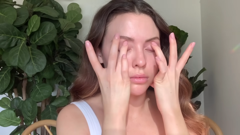 A Woman Gently Massaging Her Eyelids with Her Fingers, Sitting in A Serene Setting with Green Plants in The Background, Promoting Relaxation and Stress Relief