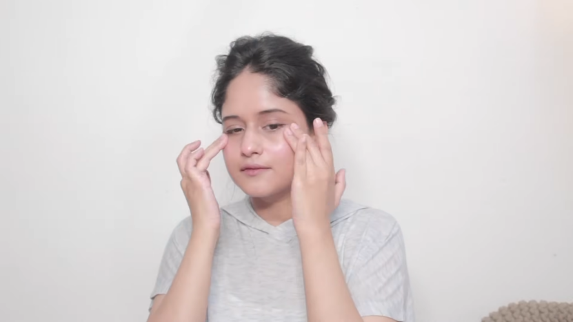 A Woman in A Light Gray Top Performing a Facial Massage by Gently Pressing Her Fingers on Her Cheekbones, Set Against a Plain White Background